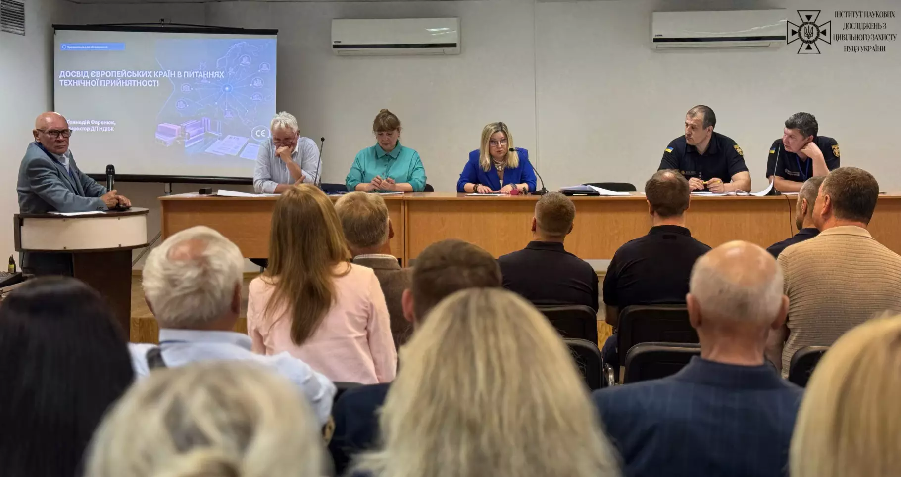 зображено конференцію, де президія з п'яти осіб за довгим столом та чоловік за трибуною виступають перед аудиторією, а на екрані позаду демонструється презентація на тему європейського досвіду технічної придатності.