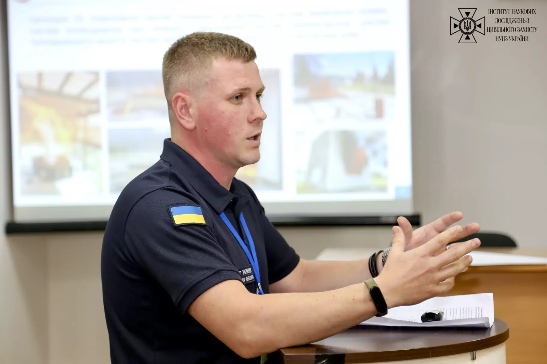 ображено молодого чоловіка у формі рятувальника ДСНС України з прапором на рукаві, який виступає за трибуною та активно жестикулює руками на тлі екрана з презентацією.