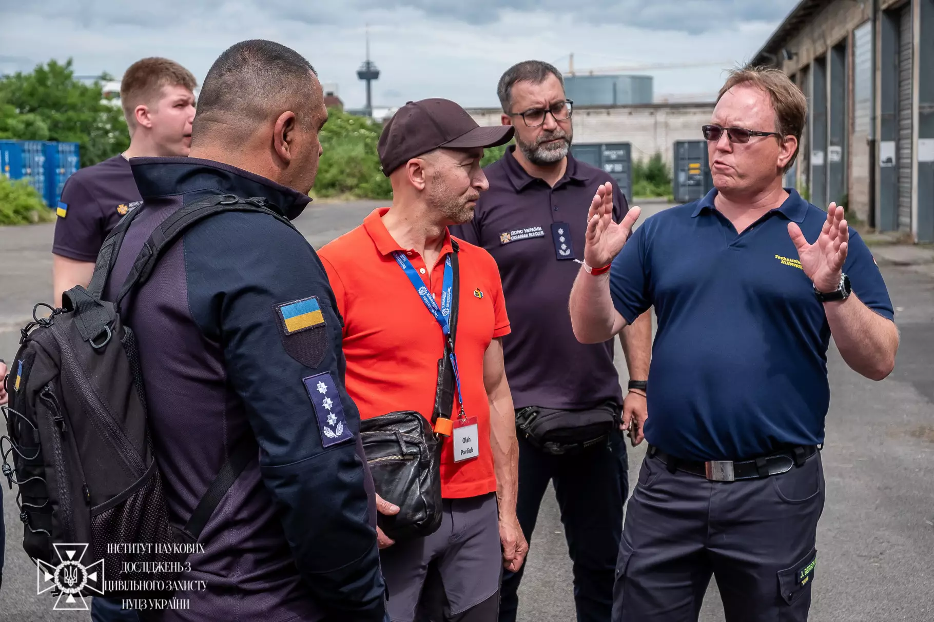 Група чоловіків, серед яких представники ДСНС України в темній уніформі з українськими шевронами та німецькі колеги, стоять просто неба на території промислового або навчального об'єкта й жваво спілкуються. Чоловік праворуч у синій футболціполо активно жестикулює, пояснюючи щось присутнім. У лівому нижньому кутку фото розміщено білий логотип Інституту наукових досліджень з цивільного захисту НУЦЗ України.