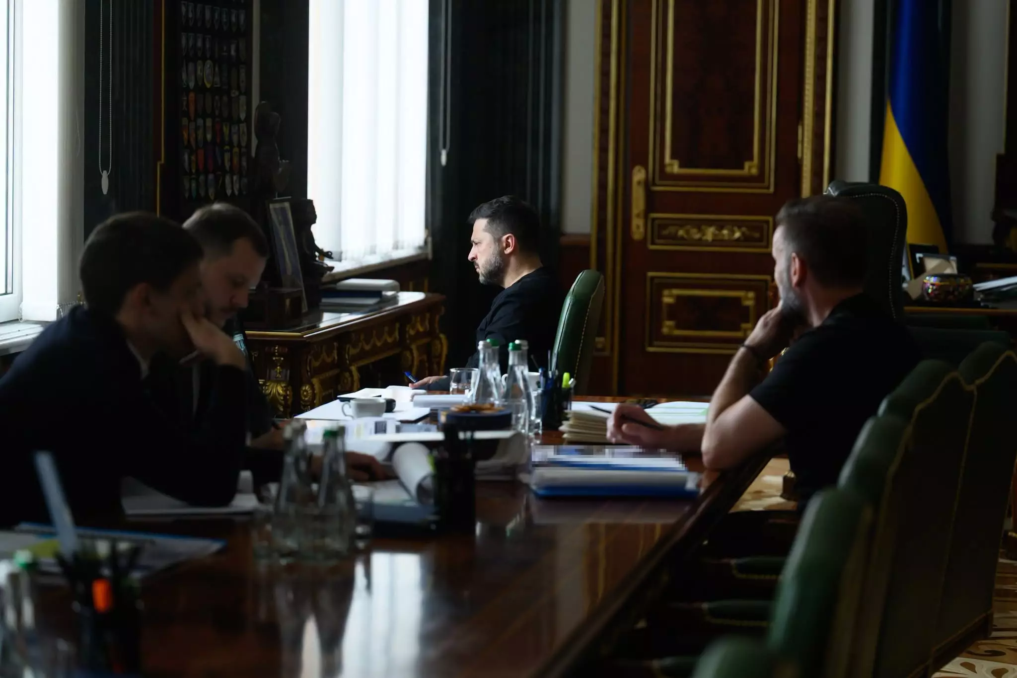 Володимир Зеленський проводить зустріч у Кабінеті Президента. Він сидить у центрі столу, обличчям до вікна, тримаючи ручку. Два чиновники сидять ліворуч, а один — праворуч, усі заглиблені в обговорення. На столі багато паперів та пляшки з водою. За ними, біля стіни, видно колекцію нагород та вікна. Праворуч — український прапор.